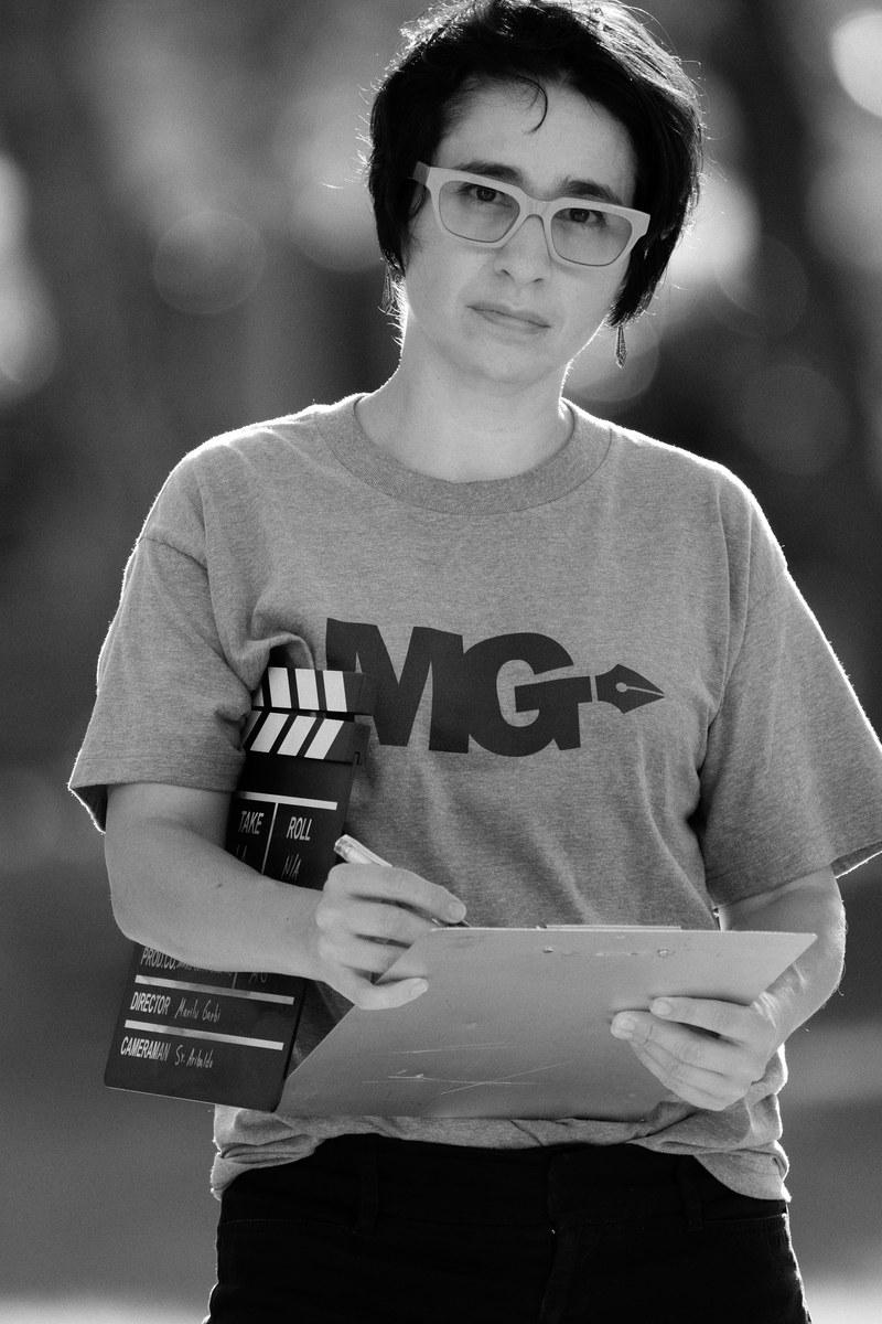 Marilu Garbi on set — holding a clapperboard and clipboard, wearing an MG-branded t-shirt. Black-and-white portrait by Harry De Zitter.
